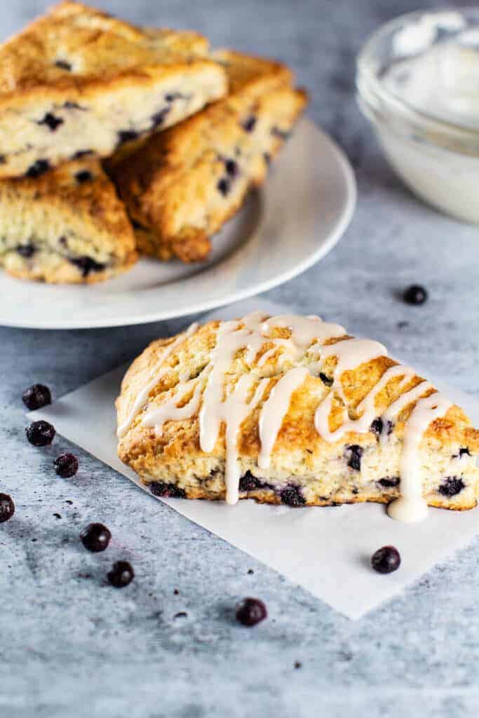 A fully glazed blueberry scone with a plate full of scones and blueberries scattered throughout