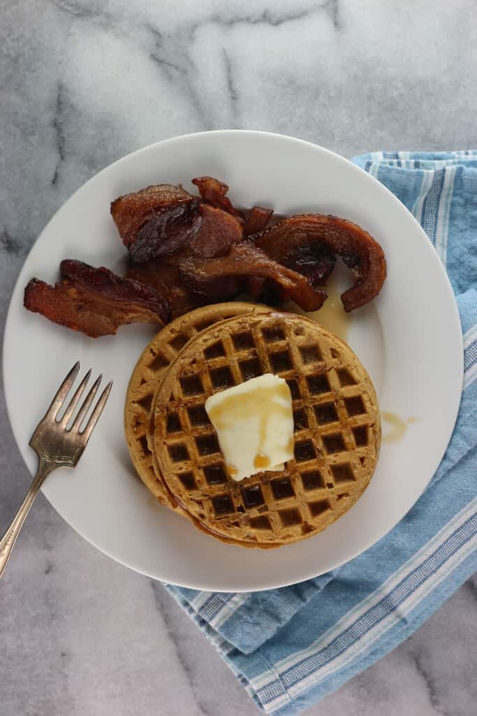 Fully heated Trader Joe's Pumpkin Waffles with butter, syrup and a side of bacon on a white plate with a blue napkin
