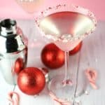 Two Holly Jolly Peppermint Martinis next to a cocktail shaker, red ornaments, and a scattering of candy canes