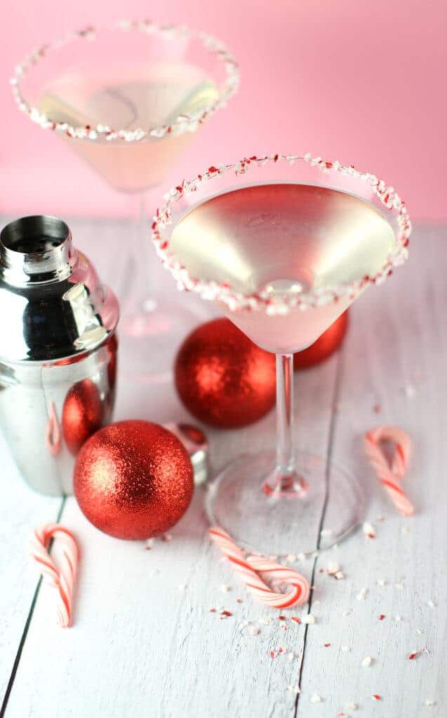 Two Holly Jolly Peppermint Martinis next to a cocktail shaker, red ornaments, and a scattering of candy canes