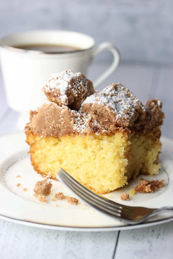 A slice of crumb cake with powdered sugar and a coffee cup in the background