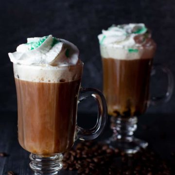 Two Irish Coffees on a dark surface surrounded by coffee beans and topped with whipped cream