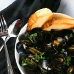 A white bowl full of Mussels in White Wine Sauce served with bread with a spoon and fork on a black napkin