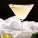 A vodka gimlet served in a martini glass garnished with a lime wedge and surrounded by a white cloth and squeezed limes