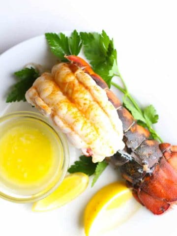A fully cooked lobster tail with lemon, parsley, and melted butter on a white plate