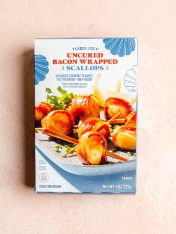 An unopened box of Trader Joe's Uncured Bacon Wrapped Scallops on a pink surface.