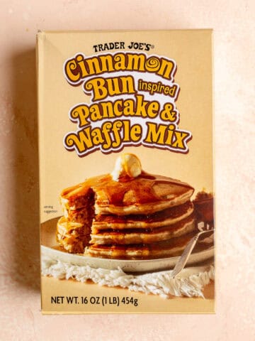 An unopened box of Trader Joe's Cinnamon Bun Inspired Pancake and Waffle Mix on a pink surface.