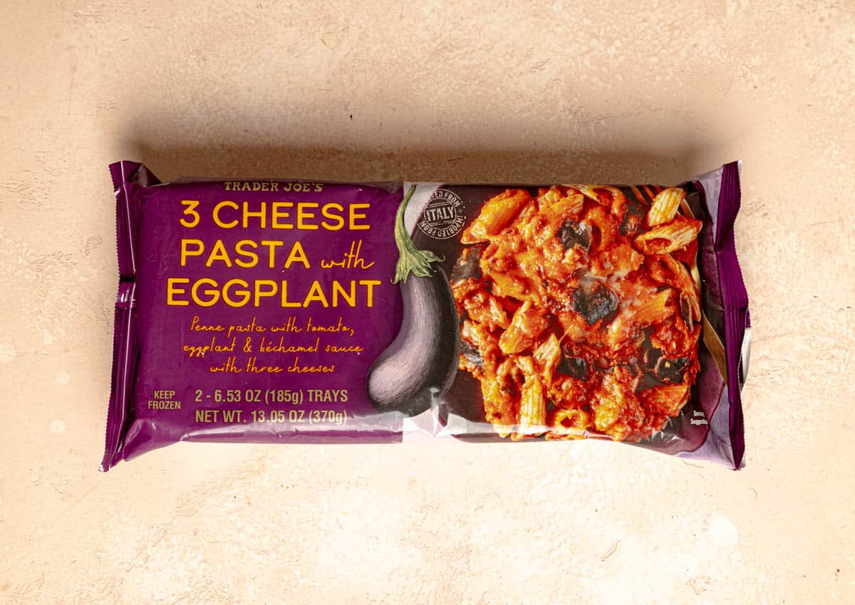 An unopened package of Trader Joe's 3 Cheese Pasta with Eggplant on a pink surface.