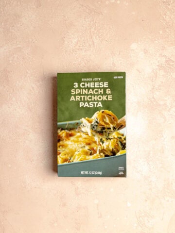 An unopened box of Trader Joe's 3 Cheese Spinach and Artichoke Pasta on a pink surface.