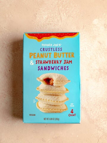 An unopened box of Trader Joe's Crustless Peanut Butter and Strawberry Jam Sandwiches on a pink surface.