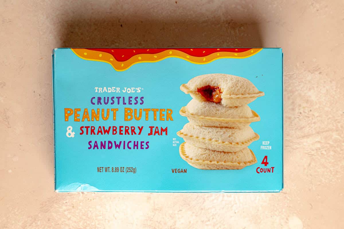 An unopened box of Trader Joe's Crustless Peanut Butter and Strawberry Jam Sandwiches on a pink surface.