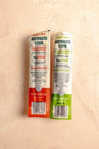 Two unopened Trader Joe's Antipasto Sticks on a pink surface.