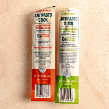 Two unopened Trader Joe's Antipasto Sticks on a pink surface.
