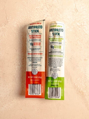 Two unopened Trader Joe's Antipasto Sticks on a pink surface.