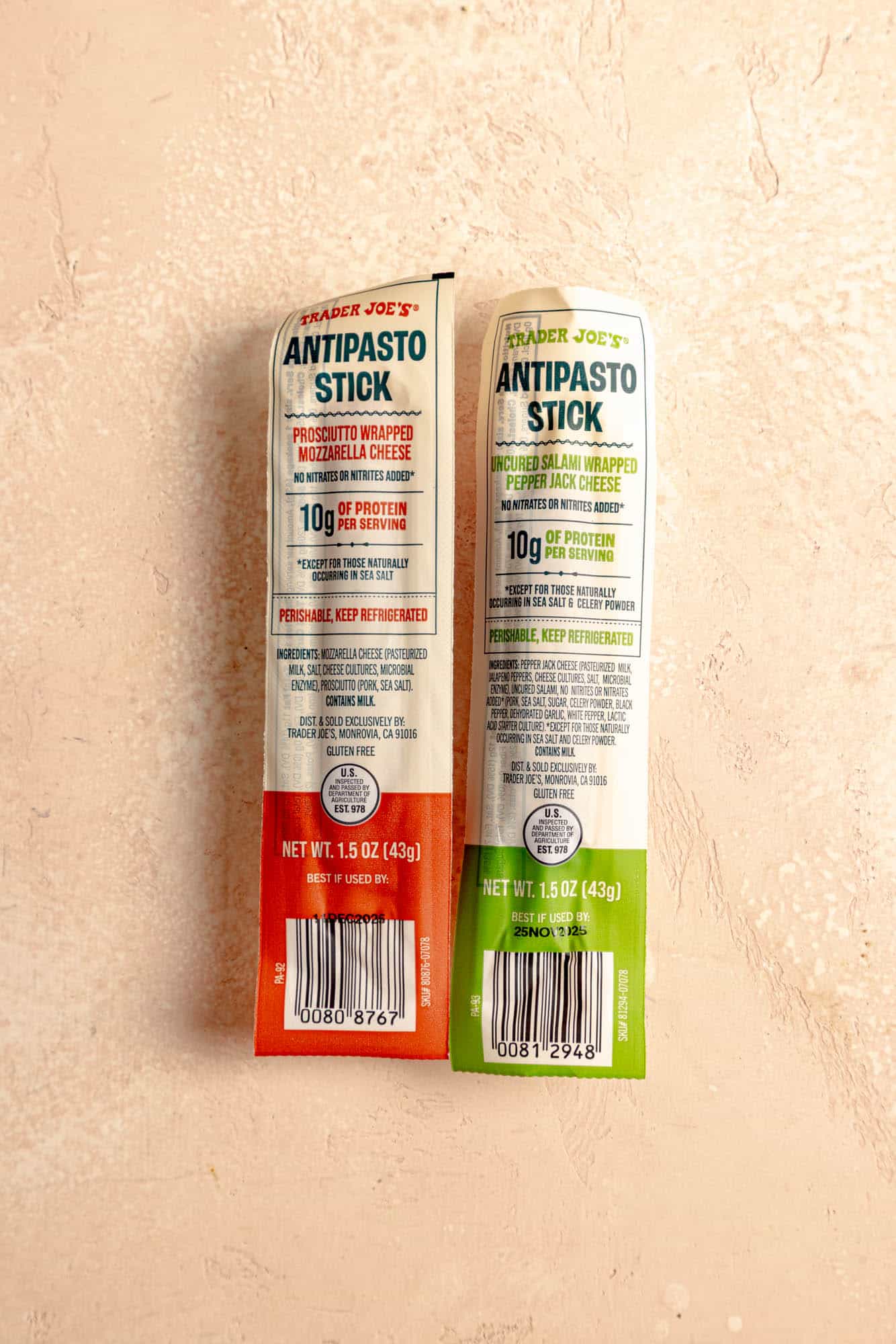 Two unopened Trader Joe's Antipasto Sticks on a pink surface.