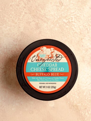 An unopened container of Trader Joe's Unexpected Cheddar Cheese Spread Buffalo Blue on a pink surface.