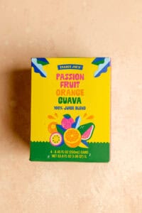 An unopened box of Trader Joe's Passion Fruit Orange Guava Juice on a pink surface.