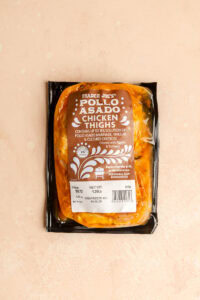 An unopened package of Trader Joe's Pollo Asado Chicken Thighs on a pink surface.