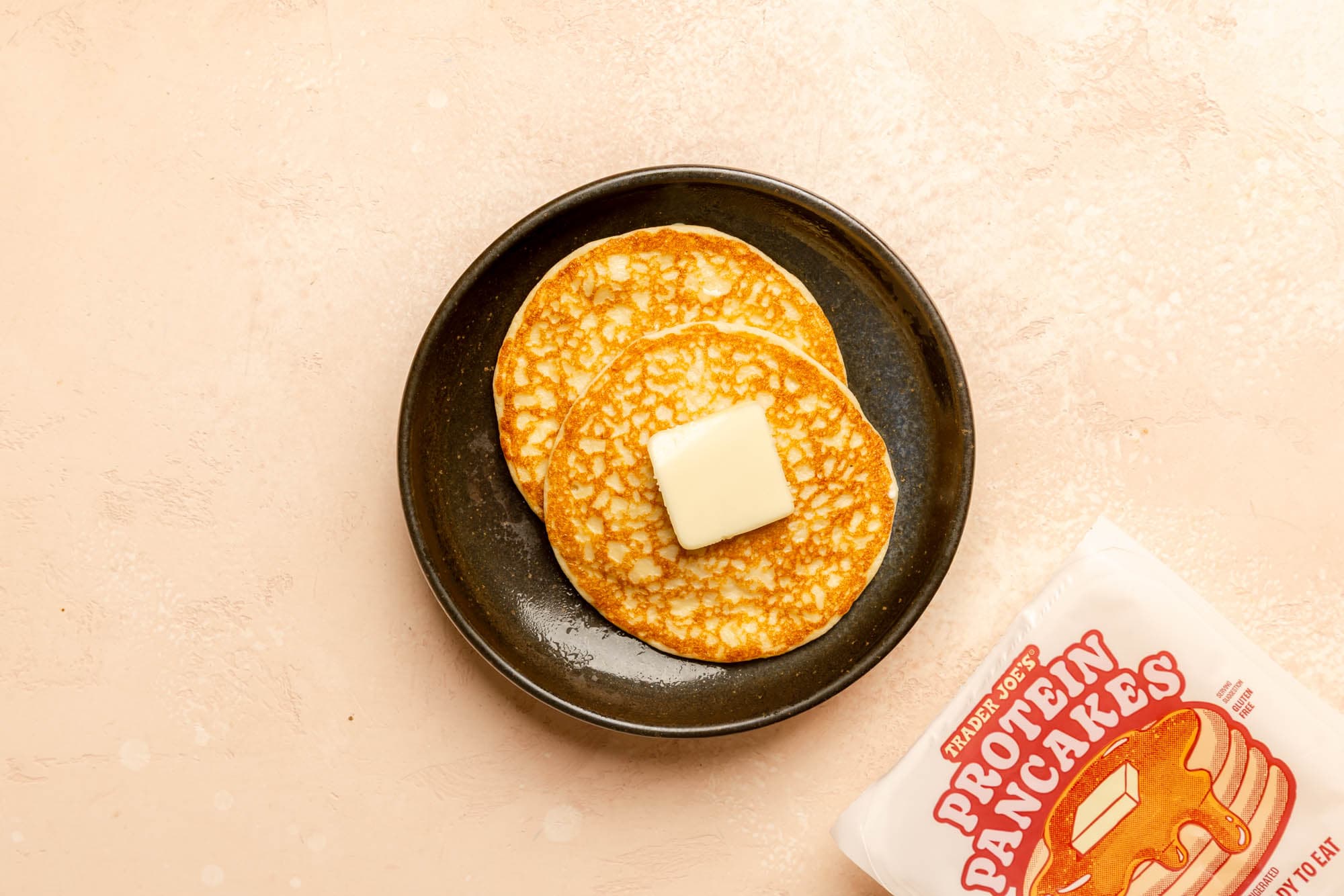 Two of Trader Joe's Protein Pancakes on a plate with butter on the top next to the original packaging.